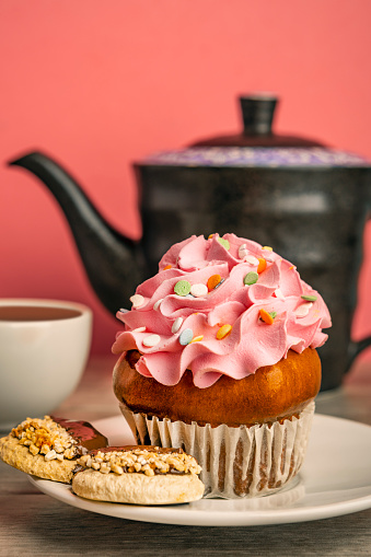 A close up of a plate with cookies and a cupcake with a teapot and tea in the background.