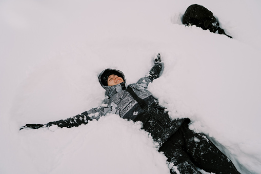 Young smiling woman in a ski suit lies in the snow with her arms outstretched near a black dog. High quality photo