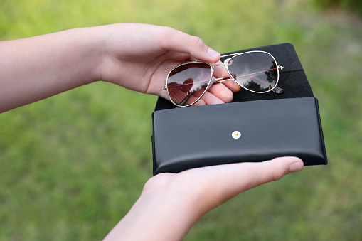 Woman putting stylish sunglasses into black case outdoors, closeup