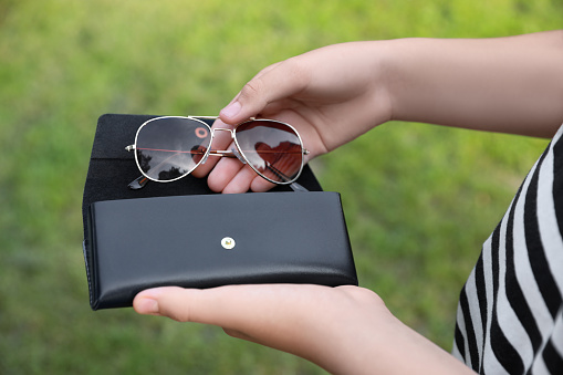 Woman putting stylish sunglasses into black case outdoors, closeup