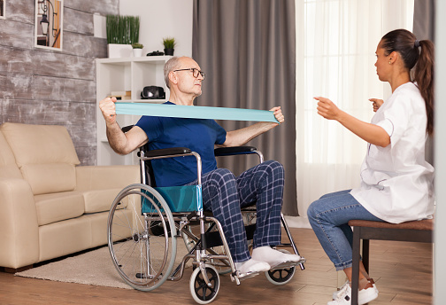 Invalid senior man in wheelchair training for muscle injury with therapist using resistance band. Disabled handicapped old person with social worker in recovery support therapy physiotherapy healthcare system nursing retirement home
