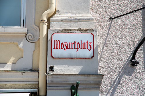 A white metal sign with the inscription Mozartplatz in old German lettering. The sign hangs on a house wall and has red letters and a red border.