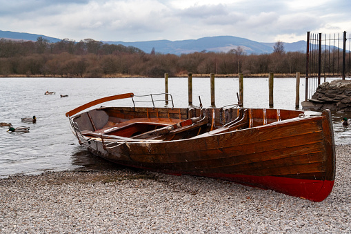 Rowing boats on the water's edge of a tranquil lake in the Lake District, set against a backdrop of lush hills and clear waters. This serene scene encapsulates the peaceful beauty of the area, renowned for its natural landscapes and leisurely boating activities.