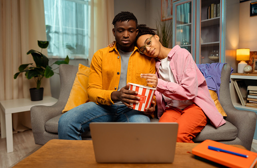 Portrait of attractive young African American couple sitting in room at home, hugging, eating popcorn, using laptop, watching video, movie. Concept of dating, relationship