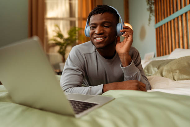 Happy smiling African American man wearing headphones lying on bed using laptop, having video call