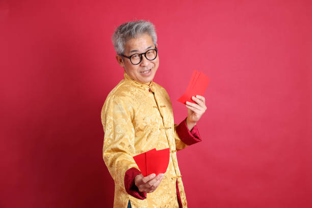 Happy Chinese new year. Asian Chinese energetic senior man wearing gold traditional cheongsam qipao or changshan dress with gesture of hand holding Chinese Red envelopes isolated on red background.