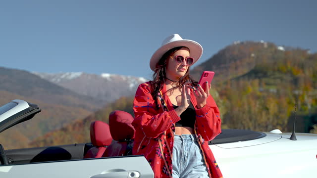 Relaxed brunette woman typing message on phone in mountains near cabriolet car.