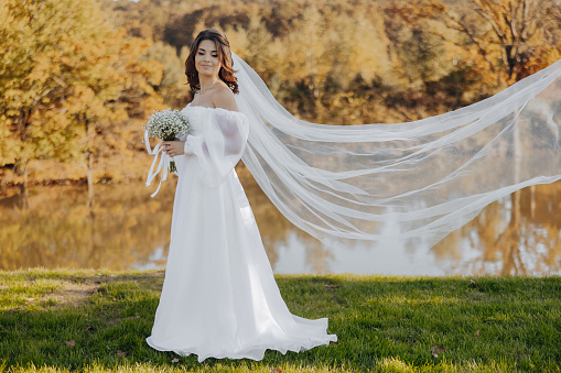 Portrait of a beautiful young bride in an elegant white dress with a long veil and a bouquet of gypsophila near the lake in soft light.