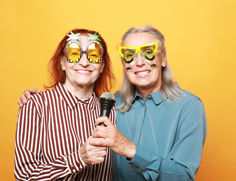 two happy elderly female friends wearing party glasses with microphone, laughs and prepares for party karaoke over yellow background