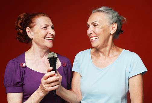 two happy elderly female friends with microphone, laughs and prepares for party karaoke over red background