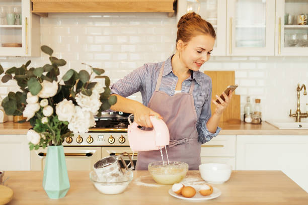 Young woman using mixer standing in kitchen and looking at recipe on mobile phone stock photo