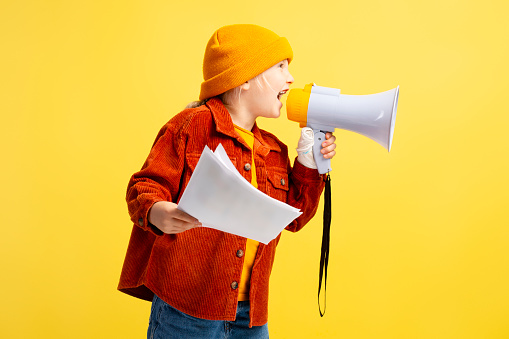Little girl wearing yellow hat, brown jacket holding paper and loudspeaker, shouting, standing isolated on yellow background. Announcement concept