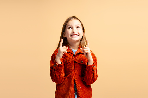 Smiling attractive little girl wearing stylish casual clothes pointing fingers up, looking up isolated on beige background. Advertisement concept