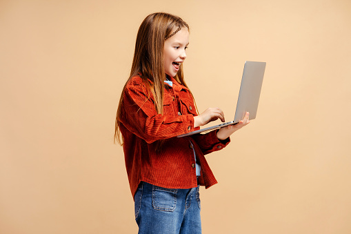 Portrait of smiling excited little girl holding using laptop, typing on keyboard, doing homework standing isolated on beige background. Technology concept