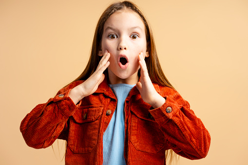 Portrait of excited shocked little girl wearing casual clothes with open mouth looking at camera isolated on beige background. Shopping concept