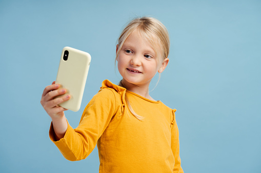 Portrait of beautiful smiling little girl, influencer holding mobile phone, taking selfie, recording video, social media, posing isolated on blue background. Technology concept