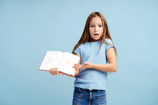 Portrait of sad, cute little girl holding notebook with bad grade, standing isolated on blue background. Back to school concept