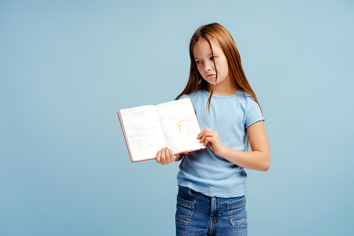 Portrait of upset little schoolgirl, student, holding notebook with bad grade, standing isolated on blue background. Back to school, progress concept