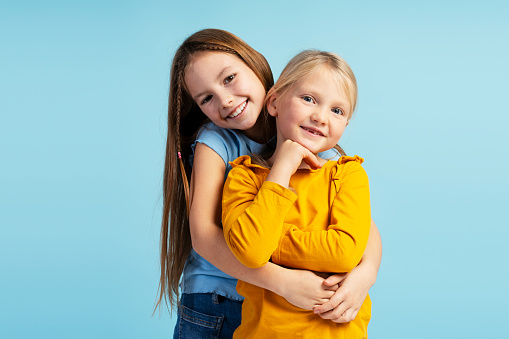 Portrait of beautiful two little sisters, hugging, looking at camera, standing on blue background. Childhood, love concept