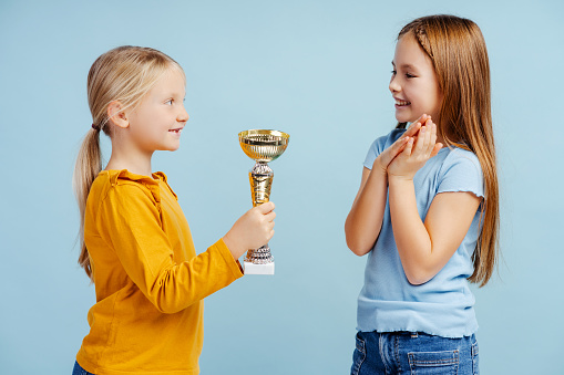 Cute little girl giving triumph cup to her sister, standing on blue background. Victory concept, successful