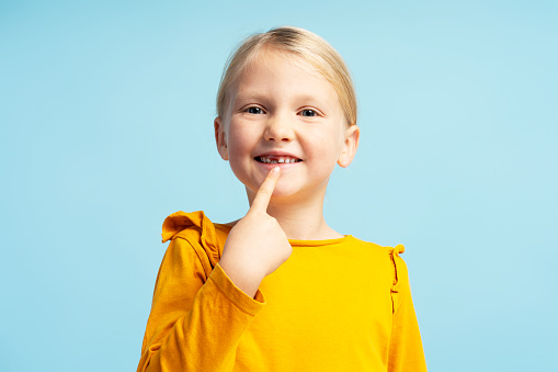 Portrait of cute little girl wearing casual clothes, pointing finger on teeth, looking at camera, standing on blue background. Dental concept, treatment