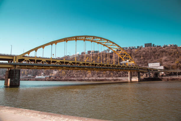 On a sunny day, the vibrant Andy Warhol Bridge stretches across the Allegheny River, framed by lush green leaves, with downtown Pittsburgh providing a picturesque backdrop.