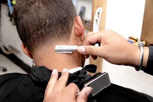 Close-up of a barber shaving the hair of a dark-skinned Latino man at the nape of his neck with a razor blade. The young man has an earring