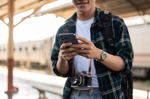 A handsome, happy young Asian man tourist backpacker using his smartphone, checking messages while standing at a platform in a railway station, waiting for his train. cropped image