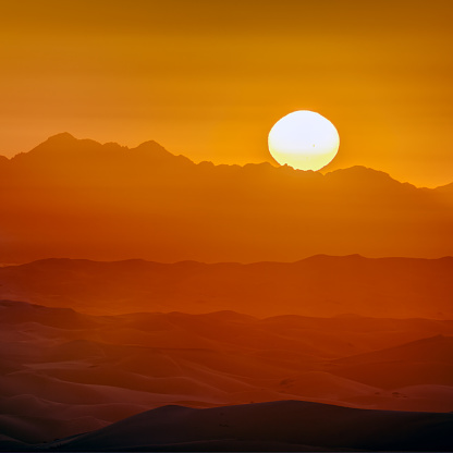 Zoomed in view of the sun rising above mountain peaks.  Golden monochrome gradients over the mountains and desert sand dunes below.