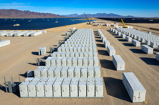 Aerial view of industrial battery units storing electricity in the desert.  In the distance are solar panels and mountains.