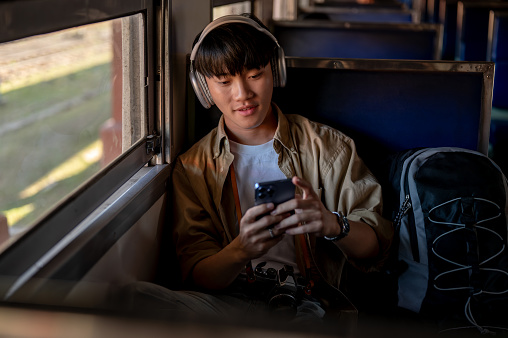 A happy and relaxed young Asian male traveler is listening to music on his headphones and using his smartphone while commuting by train to somewhere. solo travel, backpacker, vacation