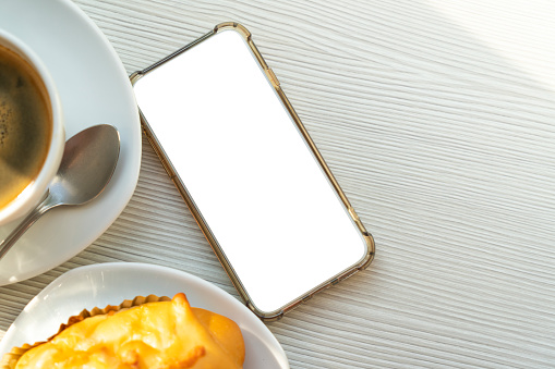 Flat lay, empty smartphone on table with hot coffee and snack or bakery in morning time. Top view image with empty space for copy and design.