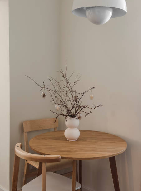 Minimalism kitchen interior - round wooden table, chairs and Easter bouquet in a ceramic vase on the table. Dry branches with glass decorative Easter eggs