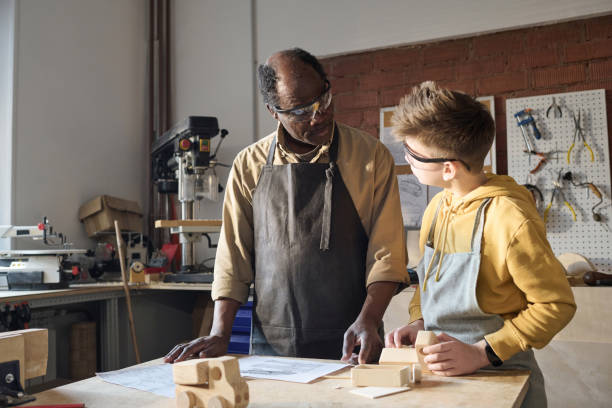 istock Black Senior Carpenter Teaching Little Boy Woodworking and Looking at Plans 1926652843