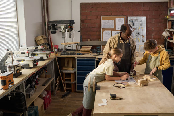 istock Two Children Building Wooden Toys with Master in Carpenting Workshop 1926656541