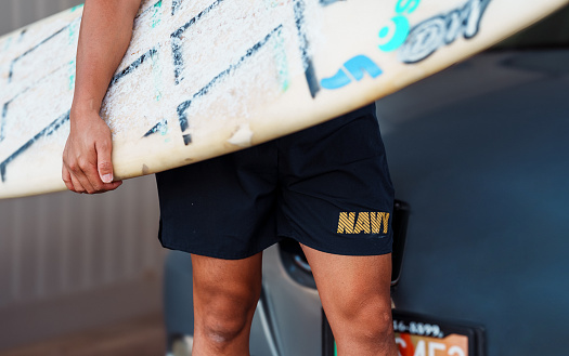 Cropped view of an unrecognizable young man of Pacific Islander descent wearing USA Navy shorts and holding a surfboard in front of his car before driving to the beach on his day off.