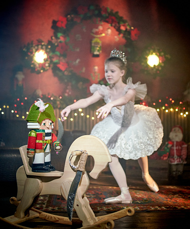 Cute pre-adolescence girl ballet dancer is standing in a ballet pose. Nutcracker is on a wooden rocking horse on foreground. Shooting in a living room with Christmas decor