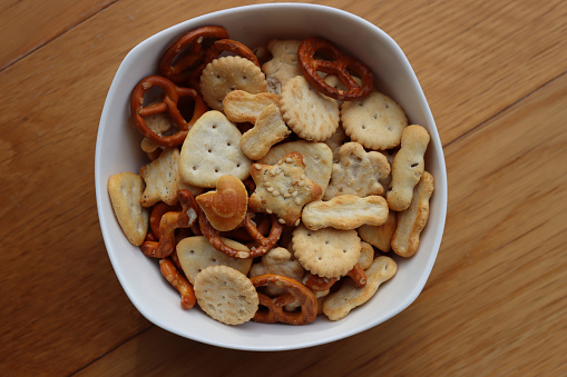 Various snack in a white bowl: pretzel with salt, crackers with seeds and herb on wooden table. Assorted party food