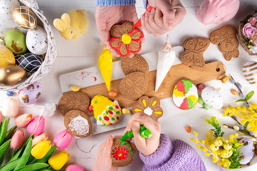 Family preparation for Easter. Holiday baking background, woman and child hands decorate homemade cookies with sugar glaze, in form of Easter symbols Easter eggs, bunny, flowers, top view copy space