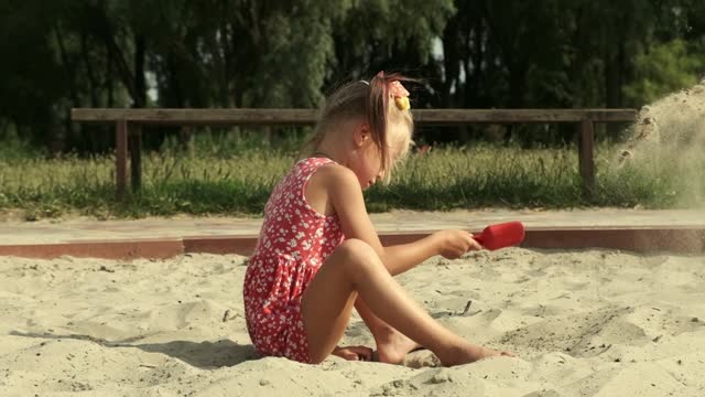 Cute little girl plays in the sandbox with a shovel wearing red dress during hot sunny summer day