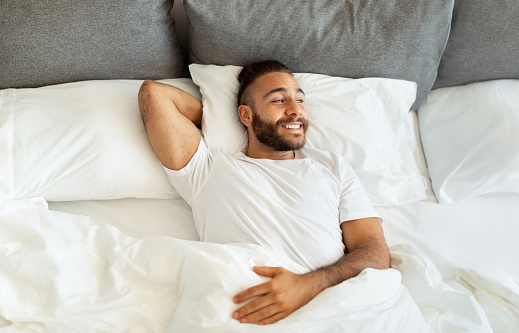 Top view of cheerful attractive bearded young guy hipster wearing white t-shirt chilling in bed in the morning at home. Positive well-rested millennial man lying in bed, look at copy space, smiling
