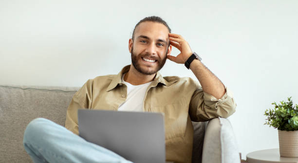 istock Smiling Millennial Man Sitting On The Couch Working On Laptop 1926849056
