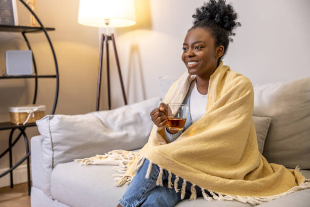 istock Smiling woman drinking tea while sitting on sofa wrapped in yellow plaid in home interior 1926860155