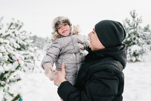 Dad hugging daughter standing in snow in winter forest. Winter holidays. Closeup. Father embracing child girl near Christmas tree in park. Happy family walking snowy mountains, spending time together.