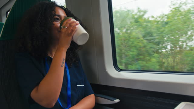Female doctor or nurse wearing uniform commuting sitting by window on moving train drinking from reusable cup - shot in slow motion