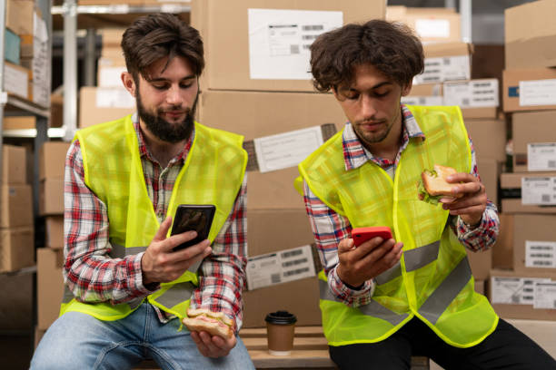istock Labor or communication concept. Happy young colleagues eating lunch and using smartphones while working in distribution warehouse 1926897490