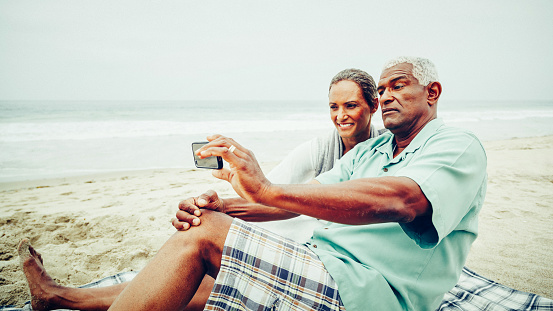 Senior African American couple having fun taking selfie with smart phone on sandy beach.