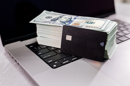 Black credit card on office desk with laptop and dollar bills. Concept for premium banking, payments, money transfers and banking services.