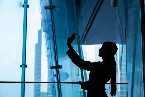 Silhouette  of young Asian woman using smart phone selfie inside building with blue tone of glass window.