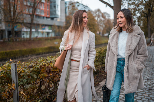 Two mid adult women, a Caucasian and an Asian, enjoying a friendly walk together, dressed in fashionable coats on an autumn day in the city.